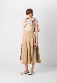 Top sans manches beige, jupe midi plissée beige, chaussures crème, pull gris clair drapé sur les épaules, lunettes de soleil rouges, sac à main rouge.