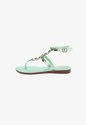 Sandal in verde menta con suola piatta, cinturini metallici e pietre decorative verdi lungo la parte superiore. Include una cinghia regolabile alla caviglia.