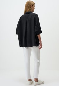 Black oversized shirt with short sleeves and a back pleat, paired with white slim-fit pants and white sneakers on a plain background.