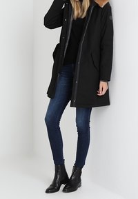 Czarna parka z futrzanym kołnierzem, zapięcie na guziki, kieszenie z przodu, w połączeniu z niebieskimi wąskimi dżinsami i czarnymi botkami za kostkę. Gładka tekstura materiału.