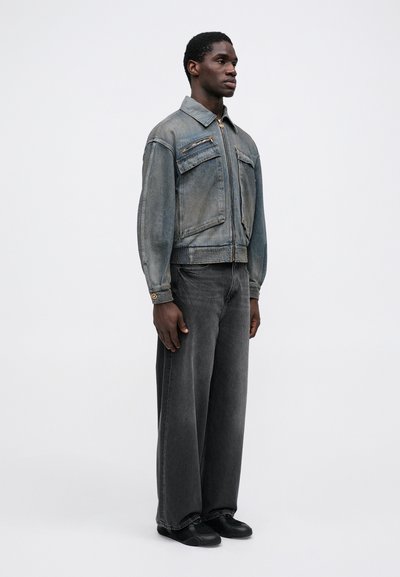 Denim jacket in faded blue-gray with zippered pockets, ribbed cuffs, and a straight silhouette. Paired with dark gray wide-leg jeans.