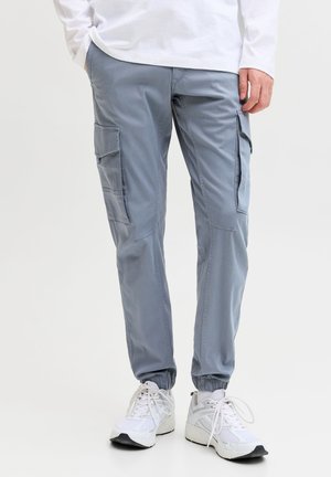 Homme portant un pantalon cargo bleu clair avec des chevilles élastiques et des baskets blanches, debout avec une main dans la poche sur fond blanc.