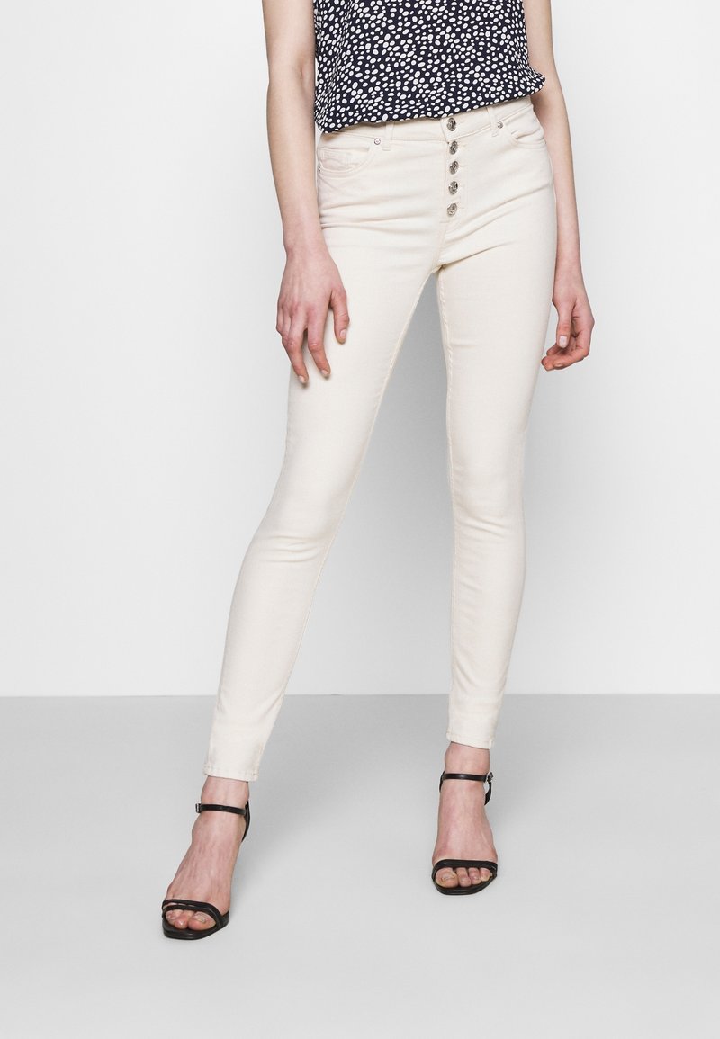 Jean skinny crème à taille haute, doté d'une fermeture à cinq boutons et de poches avant. Associé à un top à pois et des sandales noires à talons.