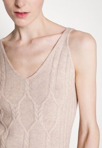 Canotta lavorata a maglia beige chiaro con scollo a V e motivo a treccia testurizzato, con una forma aderente e dettagli a coste lungo il tessuto.
