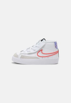 Basket montante blanche avec le logo Nike rouge, bout en daim gris, lacets blancs et languette violette à motifs sur un fond uni.