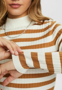 Femme ajustant la manche d'un pull côtelé à rayures marron et blanches avec un col montant et un collier en chaîne argentée.