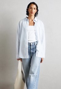 Blå och vit randig oversized skjorta med krage och knappar, bärs över en vit ribbad tanktop, ihop med högmidjade ljusblå jeans.