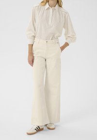 Blouse claire texturée avec col boutonné et manches bouffantes, associée à un pantalon crème à jambes larges et des baskets blanches à rayures noires.