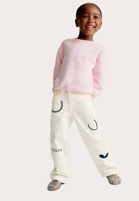 Hellrosa Sweatshirt, cremefarbene Jeans mit bestickten Mustern, mit dem Schriftzug "ALO HA" und Formen. Graue Schuhe mit strukturiertem Design.