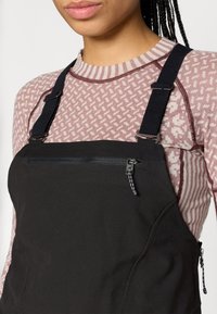 Melni sali ar regulējamiem siksnām un kabatu ar rāvējslēdzēju, uzvilkti virs rozā un baltā rakstaina krekliņa ar svītrainu apkakli.