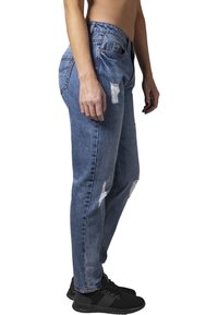 Modré džíny z denimu s volným střihem, s oprýskanými místy a roztrhanými detaily. Nosené s černými sportovními botami, zobrazené ze strany.