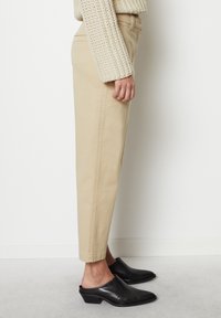 Pantaloni beige a costura corta indossati con mule con tacco in pelle nera e manica di un maglione chunky color crema visibile su uno sfondo bianco.