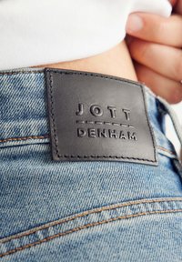 I jeans in denim presentano un tessuto blu strutturato con lievi sbiaditure. Una toppa in pelle nera con il testo in rilievo "JOTT DENHAM" è applicata sulla cintura.