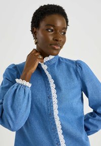 Camisa de mezclilla azul con un cuello alto de encaje blanco y mangas adornadas con encaje. Presenta cierres con botones en la parte delantera y diseños de mangas abullonadas.