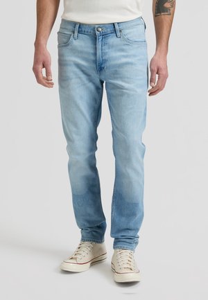 Mann trägt hellblaue Denim-Jeans und weiße High-Top-Sneaker, steht vor einem schlichten hellgrauen Hintergrund.