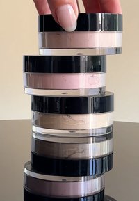 Fire gjennomsiktige krukker med laus pudder i varierende nyanser av rosa, beige og brun, hver med et svart skrulokk, stablet på en reflekterende overflate.