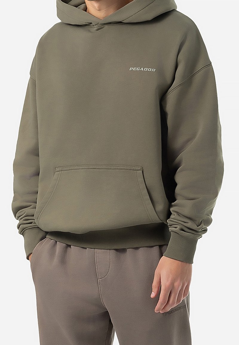 Homme portant un sweat à capuche vert olive avec poche avant et pantalon gris assorti, mains dans les poches.