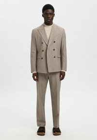 Costume double-breasted beige clair en tissu texturé, design ajusté, pantalon à devant plat et boutons foncés. Associé à des sandales noires.