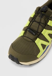 Κοντινό πλάνο αθλητικού παπουτσιού ελιάς με νέον κίτρινες επενδύσεις, μαύρο σύστημα γρήγορου δέσιμου και μπεζ σόλα σε λευκό φόντο.