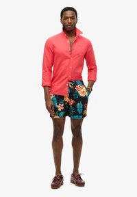 Camisa de lino color coral con diseño de botones, combinada con pantalones cortos negros con patrones multicolores de flores y hojas. Zapatos marrones.