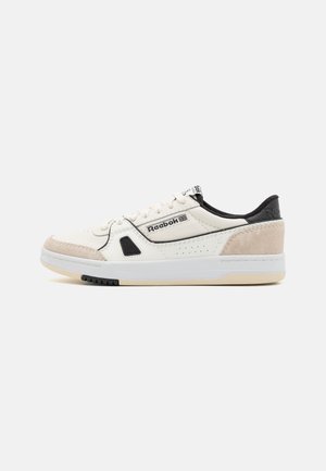 Reebok-sneakers in wit leer met beige suède accenten, zwarte afwerking, perforaties voor ademend vermogen, en een dikke witte zool met zwarte details.