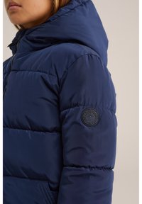 Temno modra podložena jakna s kapuco, s teksturirano tkanino, zapenjanjem na zadrgo in okroglo logotipno nalepko na rokavu.