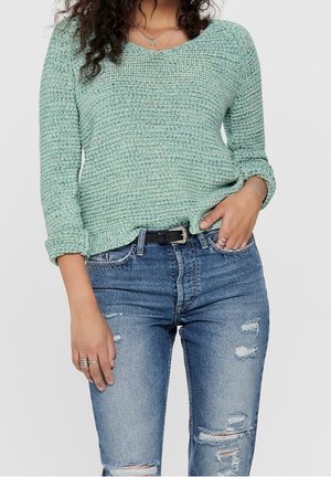 Femme portant un pull en tricot vert clair avec les manches retroussées et un jean bleu délavé avec une ceinture noire, main reposant sur la poche.
