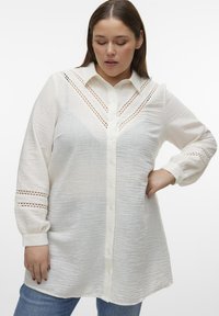 Camisa blanca ligera de botón con un acabado texturizado, que cuenta con un cuello y detalles de corte intrincados a lo largo de los hombros y las mangas.