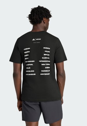 Mand iført sort t-shirt med liste over større byer og "ATHX 2026 Events"-logo på ryggen, kombineret med mørke shorts, vist bagfra.