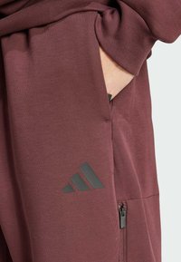 Pantalons de sport bordeaux avec une texture lisse, dotés d'une poche zippée et d'un logo Adidas gris foncé près de l'ourlet.