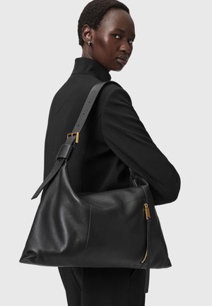 Vrouw in zwarte blazer draagt grote zwarte leren schoudertas met gouden rits en gesp, kijkt over haar schouder.