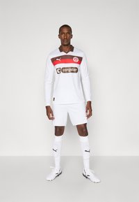 Hvit fotballtrøye med lange ermer, brune og røde detaljer, med krage og logo. Kombinert med hvite shorts og sokker, samt svarte fotballsko.
