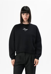 Vrouw met recht, donker haar in een zwarte sweatshirt met "Hugo" geborduurd op de voorkant en zwarte, op maat gemaakte broeken, staande tegen een witte achtergrond.
