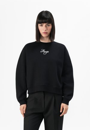 Woman with straight dark hair wearing a black sweatshirt with "Hugo" embroidered on front and black tailored trousers, standing against white background.