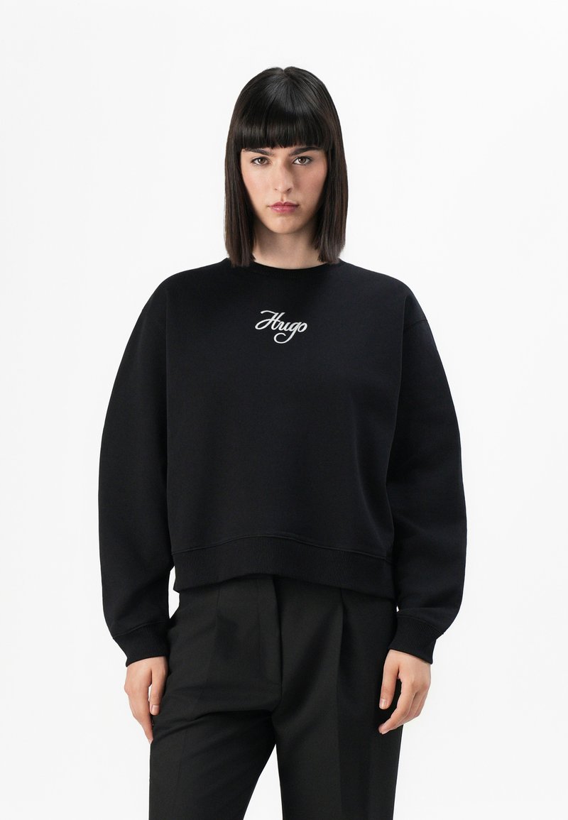 Vrouw met recht, donker haar in een zwarte sweatshirt met "Hugo" geborduurd op de voorkant en zwarte, op maat gemaakte broeken, staande tegen een witte achtergrond.