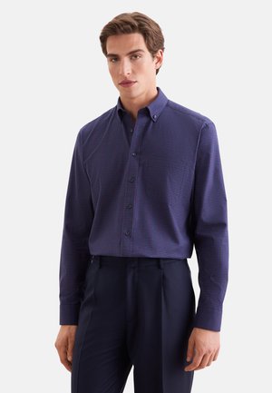 Jeune homme portant une chemise bleu foncé à manches longues boutonnée avec un petit motif à carreaux et un pantalon foncé sur un fond uni.