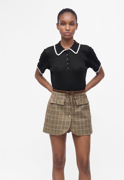 Femme debout, les mains derrière le dos, vêtue d'un haut noir à manches courtes avec un col bordé blanc et un short marron à carreaux avec poches avant et boutons.
