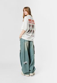 Camiseta blanca de gran tamaño con texto gráfico rojo "MOD WAVE MOVEMENT" en la espalda. Jeans holgados de mezclilla azul desgastada y zapatillas blancas gruesas.