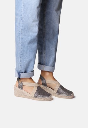 Wedge espadrilles with metallic grey fabric and beige straps, featuring an open back and woven jute sole, paired with rolled-up denim jeans.