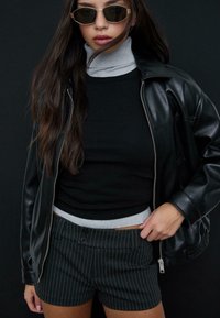 Czarna skórzana kurtka, szary golf i czarne spodenki w paski. Lustrzane okulary przeciwsłoneczne. Gładkie faktury i kontrastujące kolory.