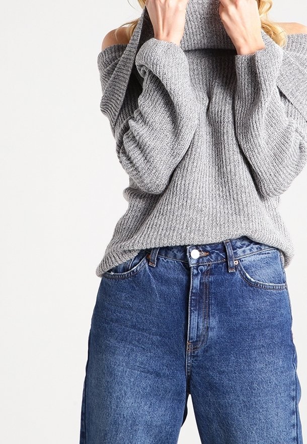 Personne portant un pull gris en maille à épaules dénudées et un jean bleu taille haute, remontant le col du pull près du visage.