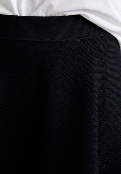 Jupe noire en tissu texturé, avec une large ceinture élastique et une forme évasée. La jupe tombe harmonieusement sans coutures visibles.