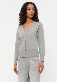 Cardigan cinza feito de malha suave com decote ondulado, fechamento com botões e barra canelada. Combinado com jeans cinza claro.