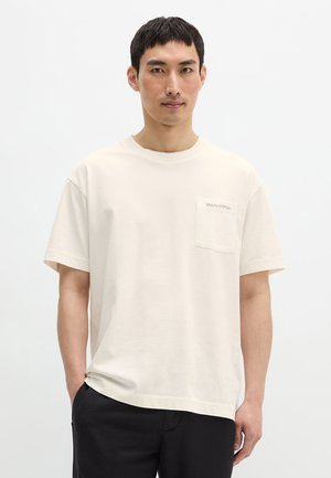 Man draagt een effen crèmekleurige korte mouwen T-shirt met een klein borstzakje en zwarte broek, staande tegen een witte achtergrond.