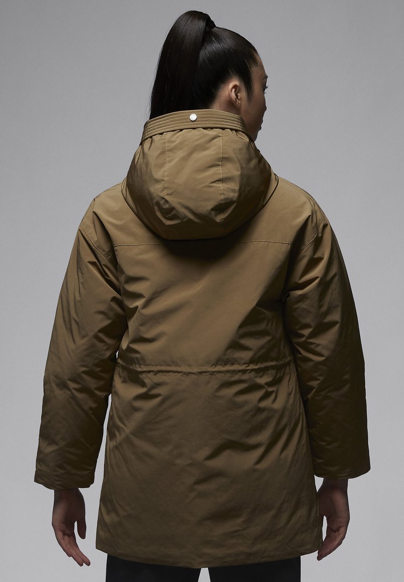 Mujer con una coleta alta que lleva un abrigo largo con capucha marrón, vista desde atrás sobre un fondo gris liso.