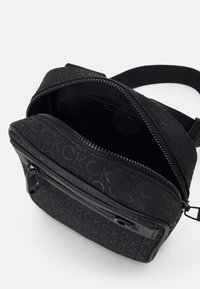 Zwarte schoudertas van gestructureerde stof met een geprofileerde hoofdcompartiment en een voorvak met rits met logo-accenten.