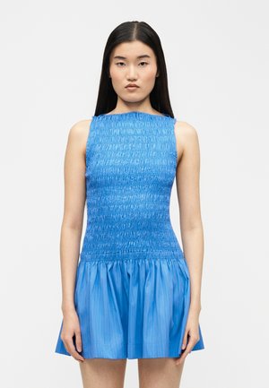 Fiatal nő hosszú, egyenes fekete hajjal, ujjatlan, kék, texturált ruhát viselve, amelynek uszálya fodros, egy egyszerű háttér előtt áll.