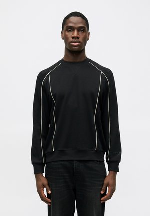 Muž stojaci čelom v čiernej mikine s bielymi detailmi a tmavých džínsoch na jednoliatom svetlom pozadí.