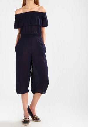 Combinaison bleu marine à épaules dénudées avec encolure volantée, taille élastique et jambes larges cropped. Associée à des chaussures à enfiler noires et à imprimé léopard.