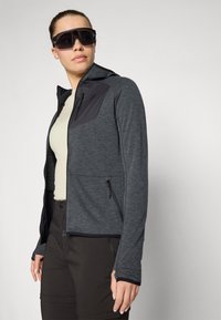 Giacca sportiva grigia con un accento nero, tasche con zip, tessuto testurizzato e cappuccio, indossata sopra una maglietta chiara e abbinata a pantaloni scuri.
